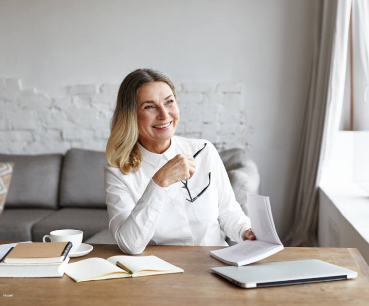 Happy, middle-aged woman sitting at desk with laptop and notepads.