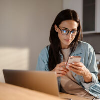 A young woman sitting in front of her laptop and looking at her smartphone.